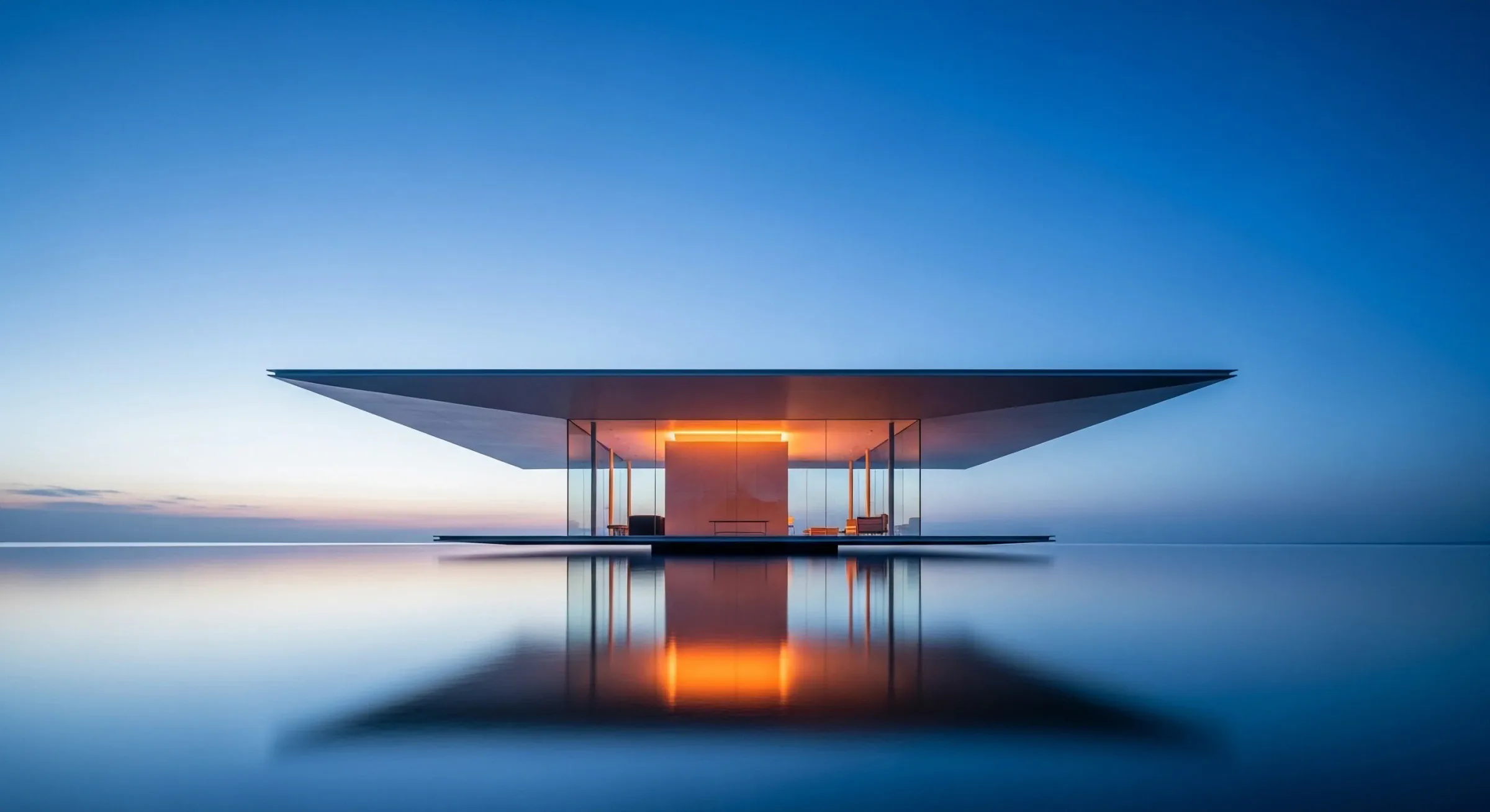 Symmetrical glass pavilion beneath a thin dark roof, illuminated at its centre and mirrored in water at blue hour.